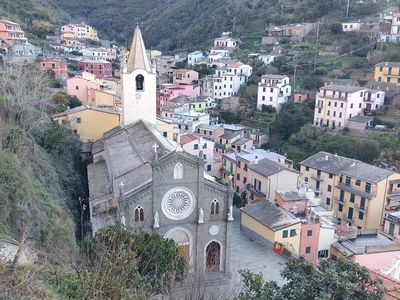 Vista panoramica della Chiesa di San Giovanni Battista e del borgo di Riomaggiore tra le case colorate e le montagne della Liguria.